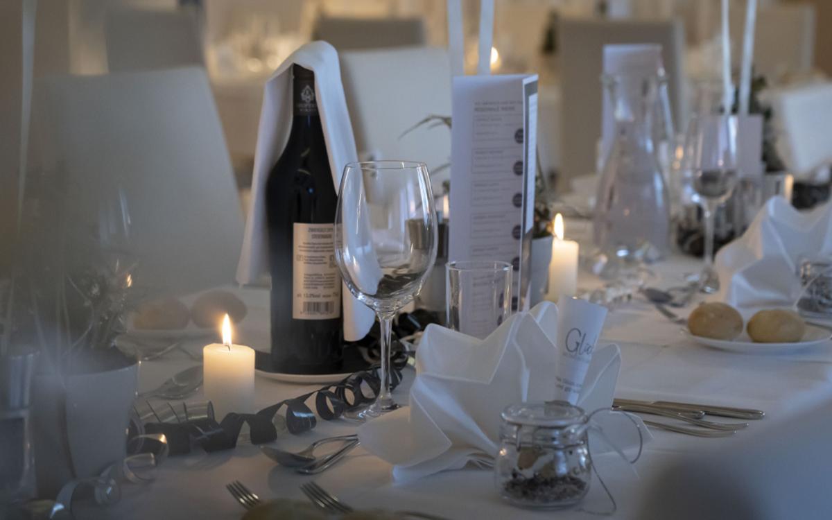 Ein weiß gedeckter Tisch für die Silvestergala mit Brötchen, Kerze, Besteck, gefalteter Serviette, einer Flasche Wein, Menüplan