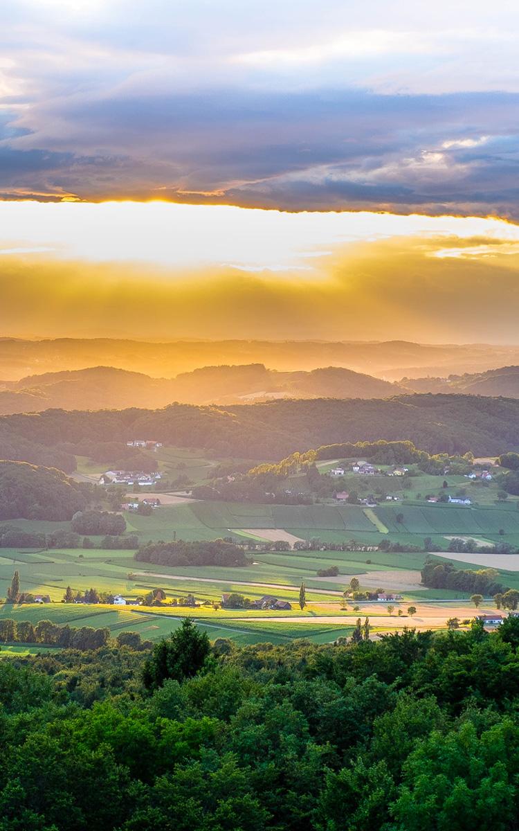Landschaftsbild - Aussicht vom Stradner Kogel bei Sonnenuntergang. Grüne Wälder, gelber Sonnenschein, Wolken am Himmer, die Sonne bricht durch Wolken heraus und am Horizont sind die Schatten viele Hügel und Berge zu sehen