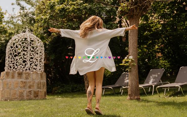 Frau in einem weißen Kleid tanzt mit ausgebreiteten Armen im Garten des Vulkanlandhotels Legenstein im Thermen- und Vulkanland Steiermark im Pride Month..