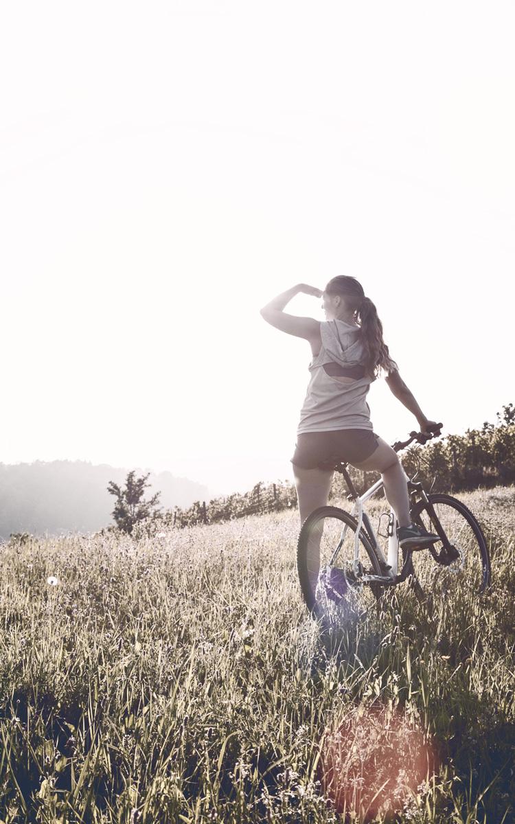 Frau steht mit Fahrrad in einer Wiese und blickt gegen die Sonne, die Hand auf der Stirn - Radfahren auf der Kraftquellentour in Bad Gleichenberg im Thermen - und Vulkanland Steiermark