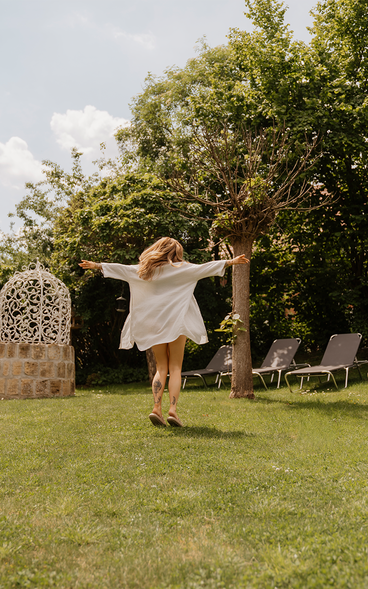 Eine Frau im weißen Kleid tanzt bei Sonnenschein durch den grünen Garten des Vulkanlandhotels Legenstein.