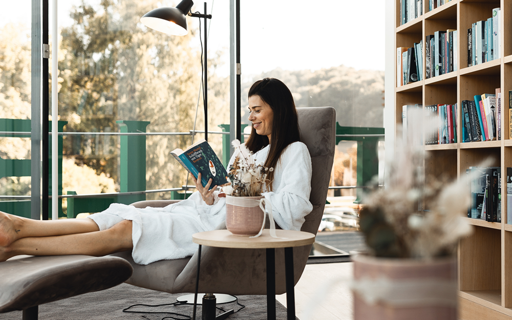 Eine Seminarteilnehmerin sitzt im Bademantel in der Bücherwurmlounge des Vulkanlandhotels Legenstein und liest ein Buch, umgeben von einer atemberaubenden Aussicht auf die hügelige Landschaft.