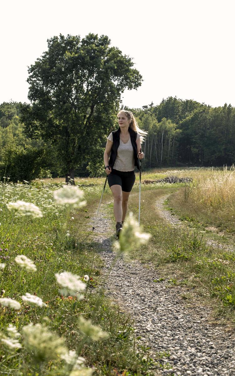 Frau walked über einen Schotterweg mit Nordic Walking - Stöcken. Davor sind weiße Blumen sichtbar, dahinter Wald.