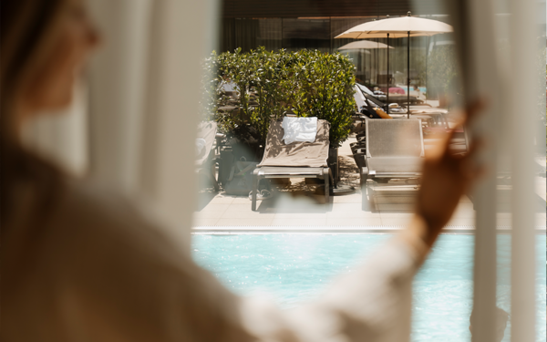 Blick durch geöffnete Jalousien auf den blauen Pool und die Sonnenliegen im Garten des Vulkanlandhotels Legenstein