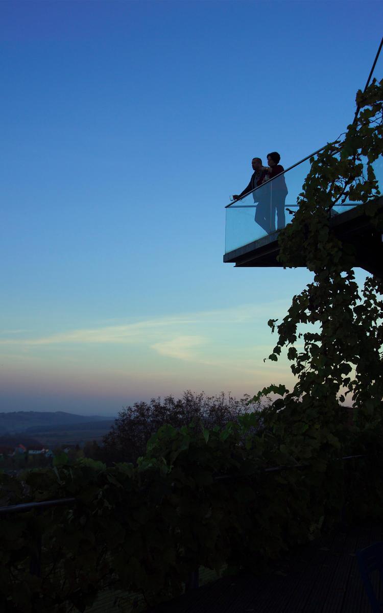 Pärchen steht auf einer Plattform oberhalb der Gesamtsteirischen Vinothek in St. Anna am Aigen und blickt bei Sonnenuntergang in die Region - Ausflugsziel vier Jahreszeiten - Bad Gleichenberg - Vulkanlandhotel Legenstein