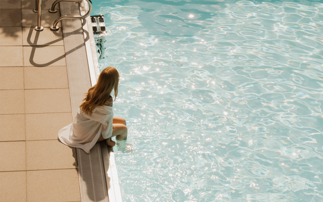 Eine Frau sitzt am Rand des glitzernden Außenpools im Vulkanlandhotel Legenstein und lässt die Füße ins Wasser hängen.