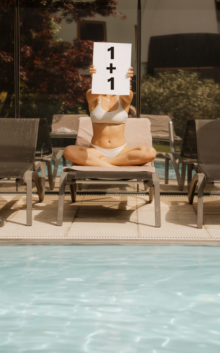 Frau im Bikini auf einer Liege am Pool hält ein Schild mit „1+1“ vor ihr Gesicht. Sommerliche Stimmung im Day Spa des Vulkanlandhotels Legenstein mit dem 1+1 Day Spa Sonderangebot.