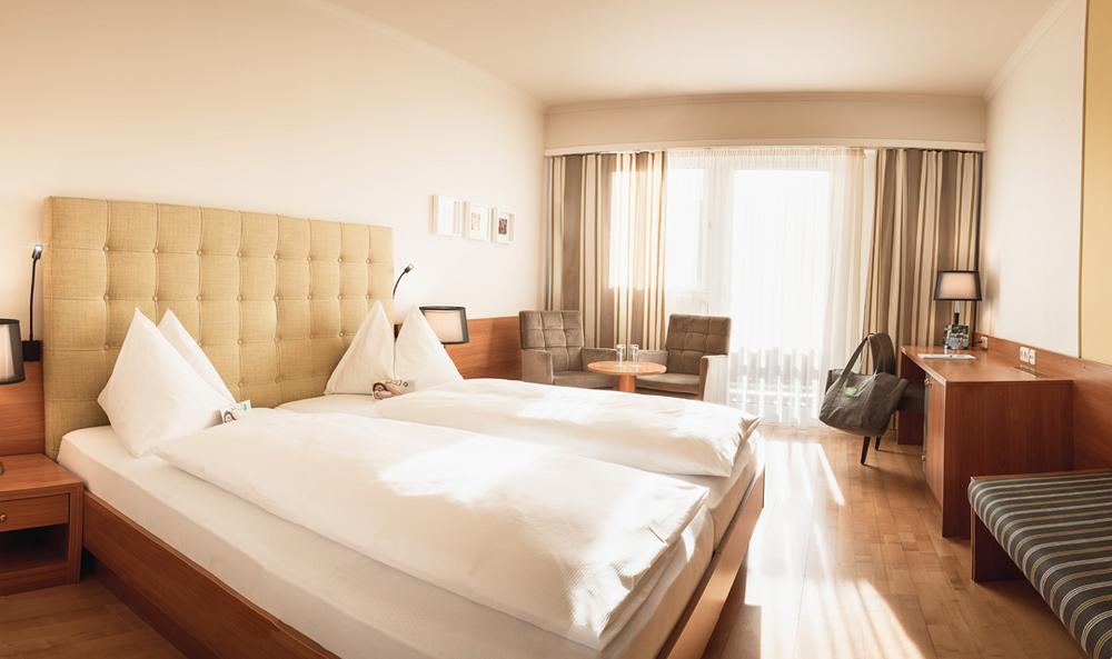 Doppelzimmer Cirkonia - Preise im Vulkanlandhotel Legenstein in Bad Gleichenberg mit Doppelbett, einem Tisch mit zwei Stühlen, eine Bank an der Wand, einem TV und einem Wandschreibtisch mit Minibar, DU und WC sind im Zimmer getrennt, Balkon mit Blick in den Garten, Klimaanlage