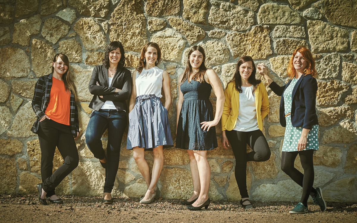 6 Winzerinnen stehen an einer Steinmauer und lächeln in die Kamera