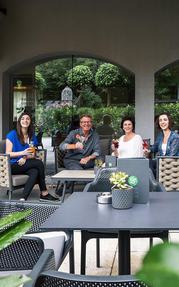Familie sitzt im Poolcafé mit Cocktails in der Hand umgeben von saftig grüner Gartenlandschaft